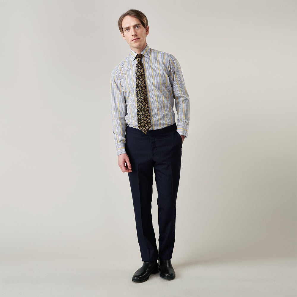 A man stands against a light background, wearing a Turnbull & Asser Yellow and Blue Fancy Check Regular Fit Shirt with T&A Collar, a patterned tie, dark dress pants, and black shoes. He faces forward with hands in pockets and a neutral expression.