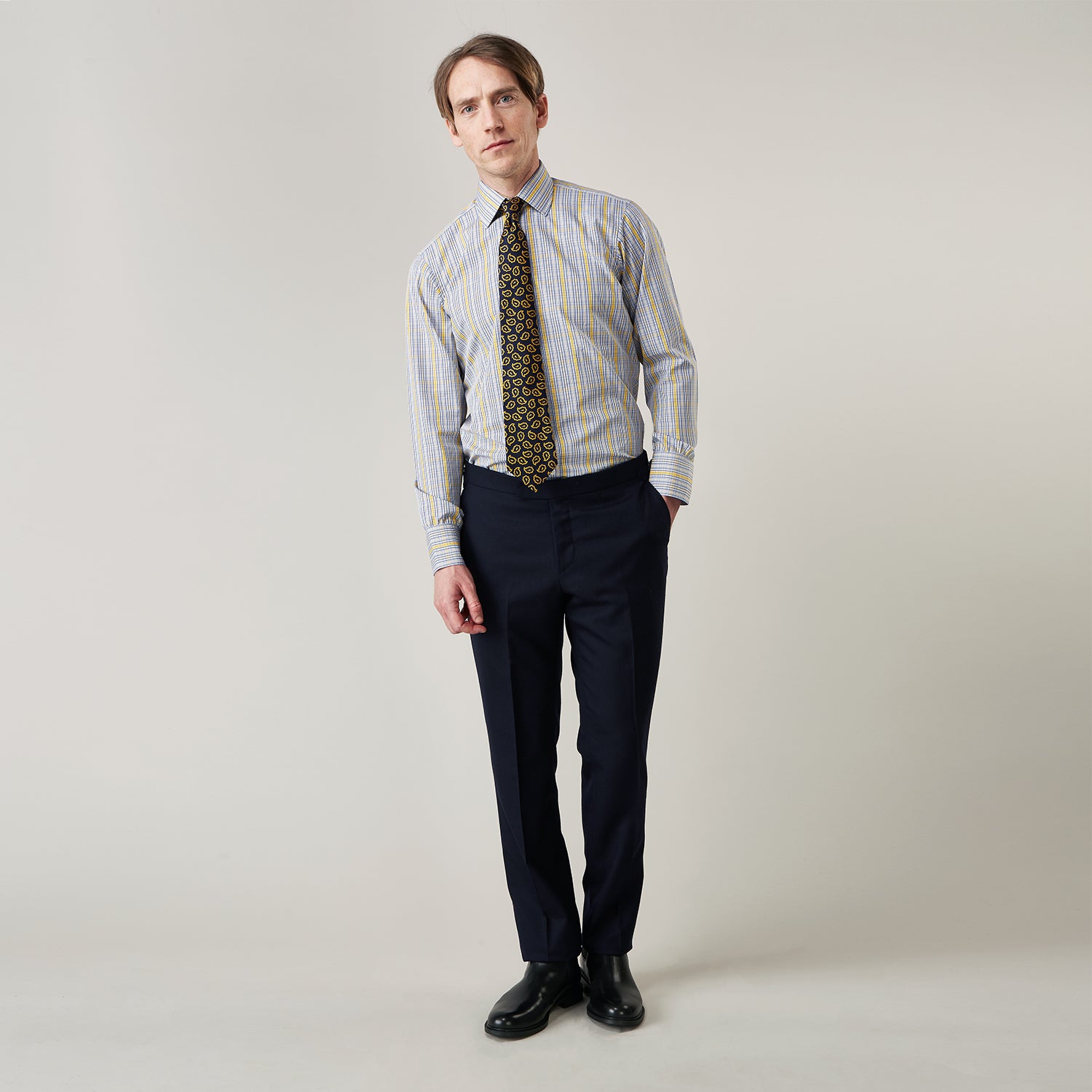 View 1 of product A man stands against a light background, wearing a Turnbull & Asser Yellow and Blue Fancy Check Regular Fit Shirt with T&A Collar, a patterned tie, dark dress pants, and black shoes. He faces forward with hands in pockets and a neutral expression.