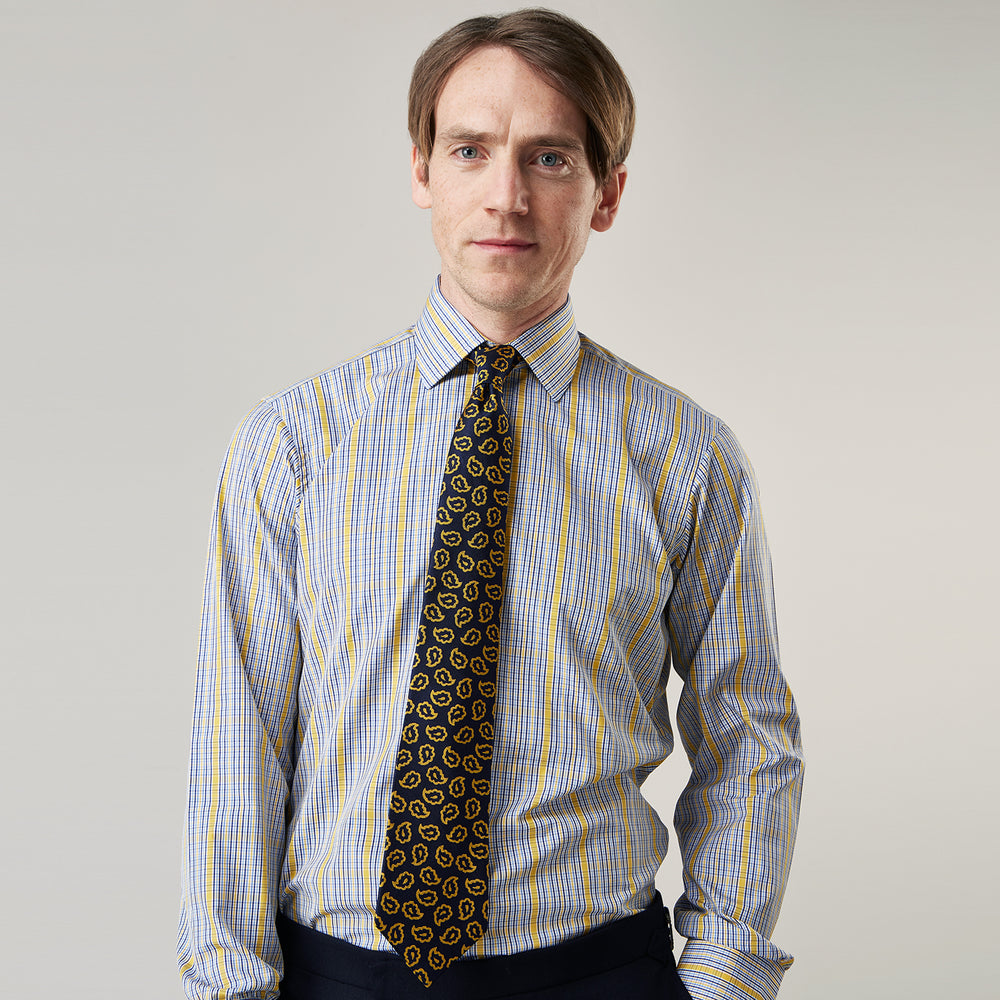 A man stands against a light background, wearing a Turnbull & Asser Yellow and Blue Fancy Check Regular Fit Shirt with T&A Collar, a patterned tie, dark dress pants, and black shoes. He faces forward with hands in pockets and a neutral expression.