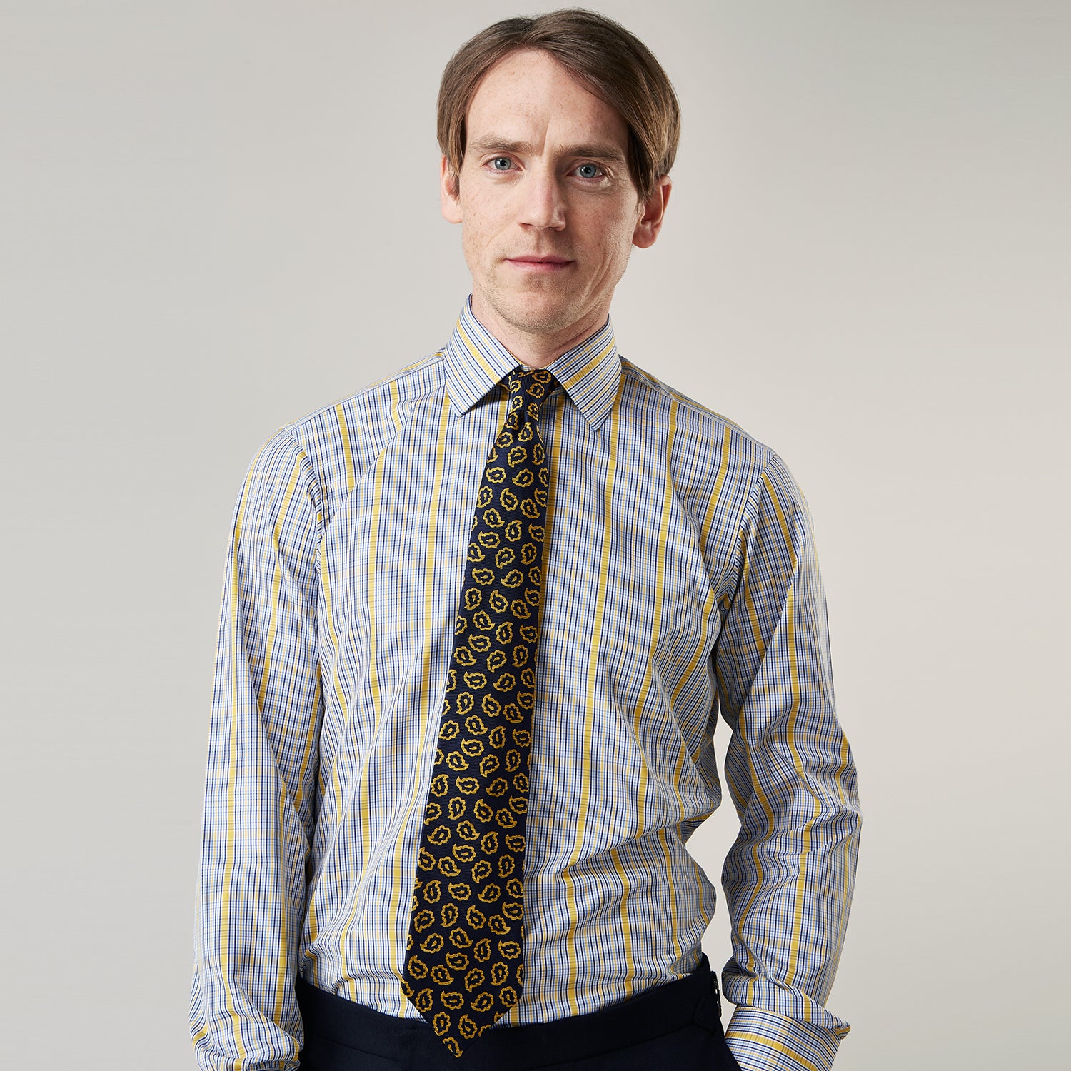 View 3 of product A man stands against a light background, wearing a Turnbull & Asser Yellow and Blue Fancy Check Regular Fit Shirt with T&A Collar, a patterned tie, dark dress pants, and black shoes. He faces forward with hands in pockets and a neutral expression.