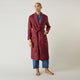 A person stands barefoot against a plain background, wearing the Turnbull & Asser Burgundy Linen Piped Gown belted at the waist over blue pajamas, looking to the side with one hand in the gown’s pocket.