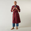 View 7 of product A person stands barefoot against a plain background, wearing the Turnbull & Asser Burgundy Linen Piped Gown belted at the waist over blue pajamas, looking to the side with one hand in the gown’s pocket.