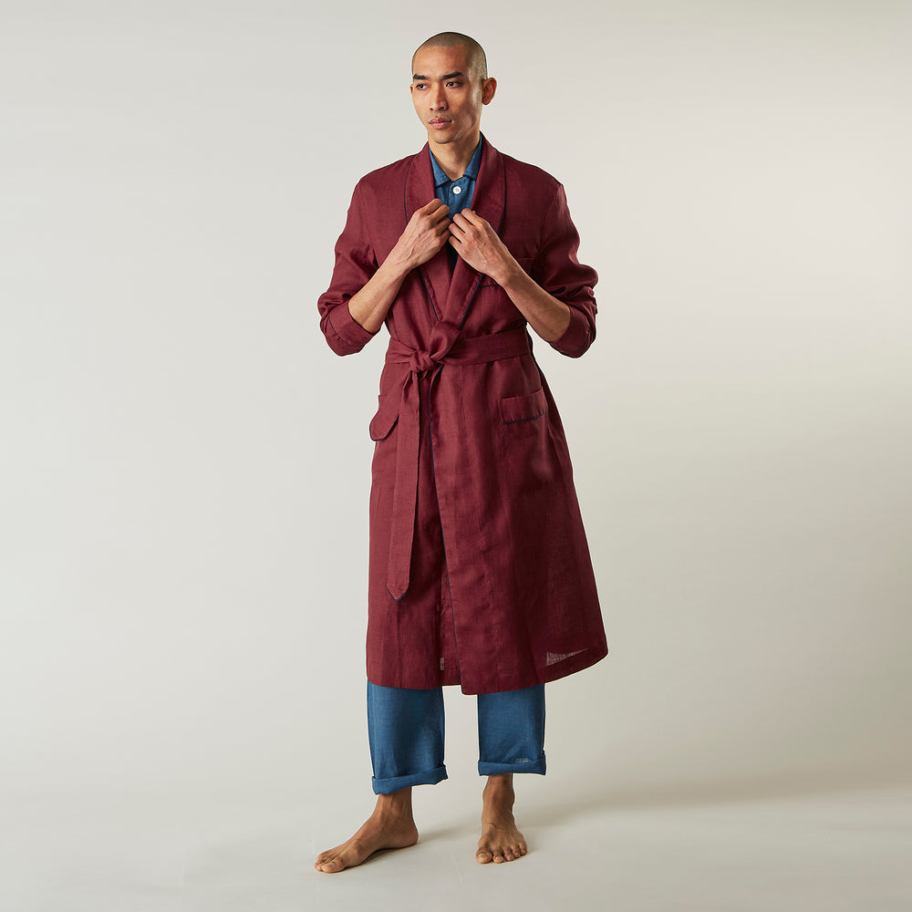 A person stands barefoot against a plain background, wearing the Turnbull & Asser Burgundy Linen Piped Gown belted at the waist over blue pajamas, looking to the side with one hand in the gown’s pocket.