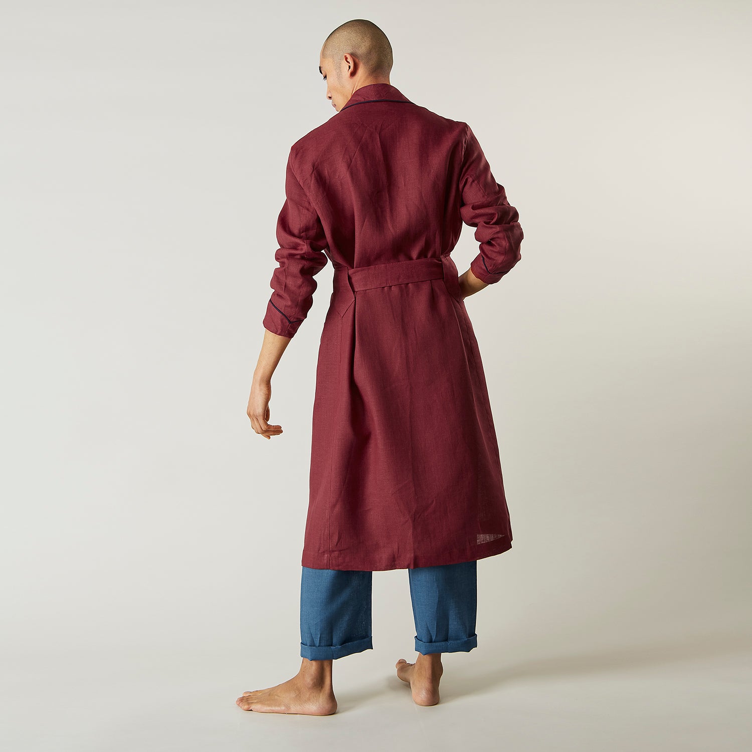 View 8 of product A person stands barefoot against a plain background, wearing the Turnbull & Asser Burgundy Linen Piped Gown belted at the waist over blue pajamas, looking to the side with one hand in the gown’s pocket.