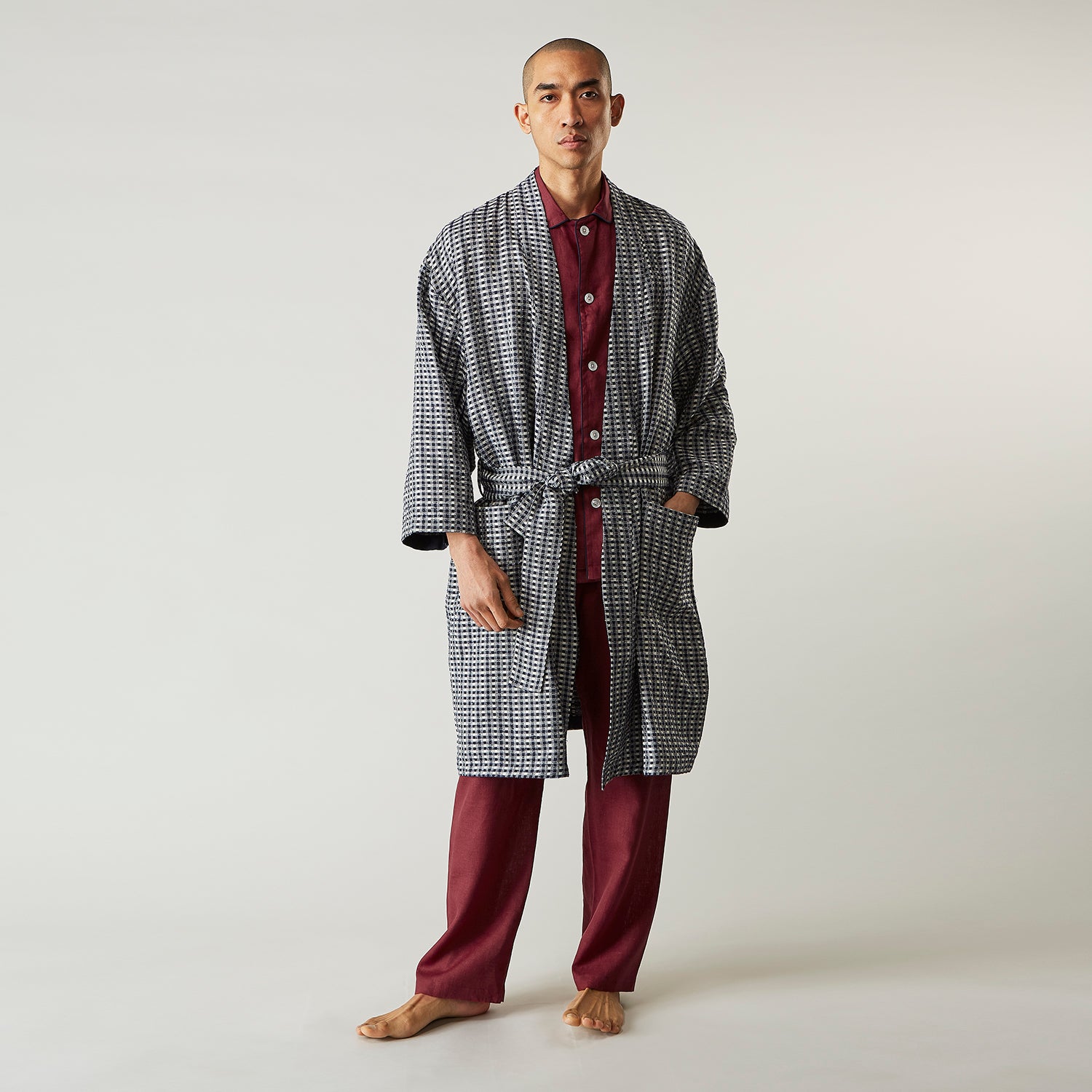 View 1 of product A barefoot person wears Turnbull & Asser's Navy Silk Chequered Kimono Gown with a belt over maroon pyjama sets, standing against a plain light background.