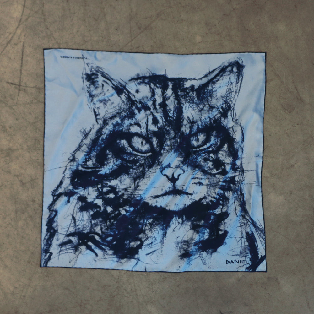 The Turnbull & Asser DANIEL PERKIN BLUE WILDCAT POCKET SQUARE features a blue-toned sketch of a tabby cat’s face with vivid eyes and fine fur detail on a pale blue background, finished with “DANISL” in the lower right corner.