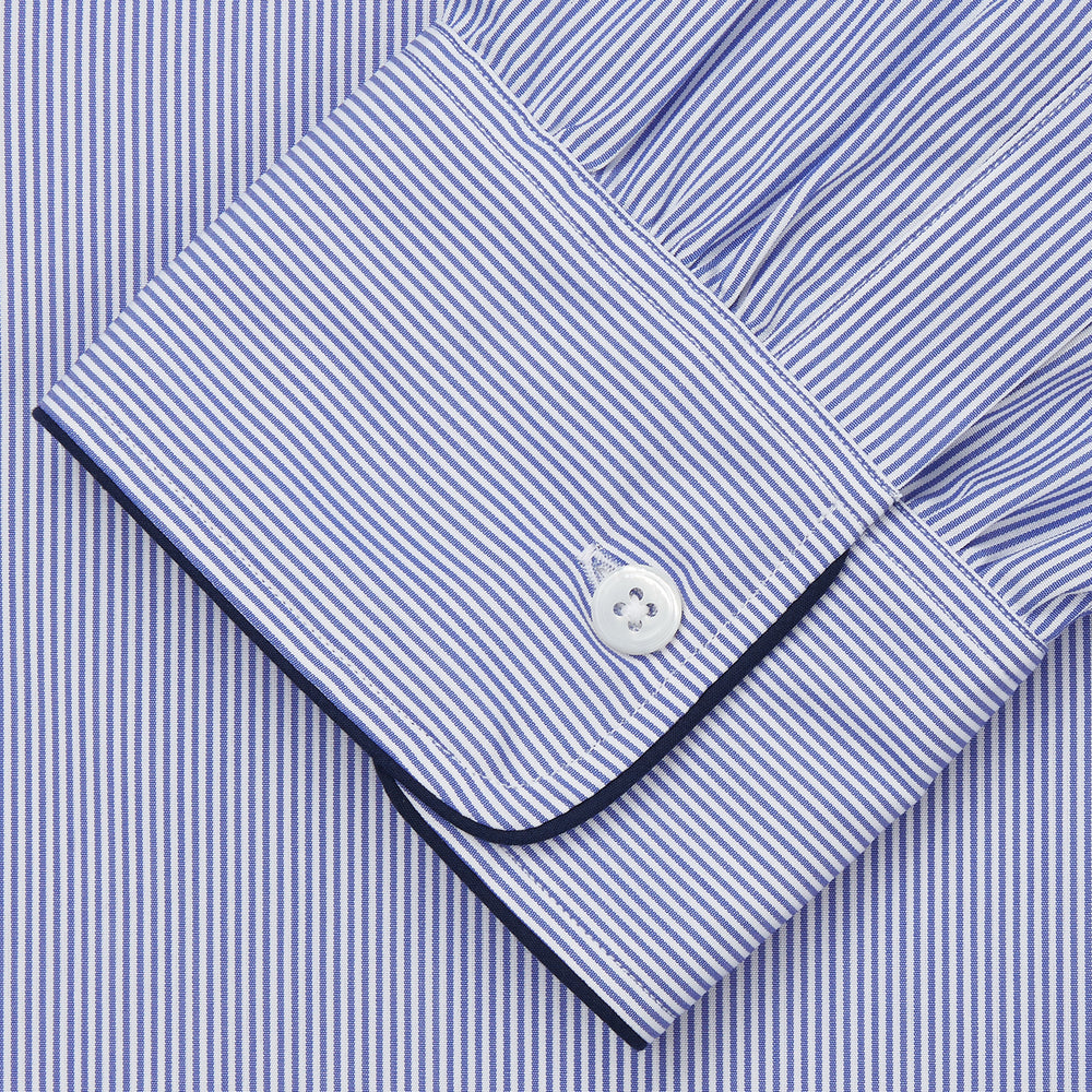 The Turnbull & Asser Blue Fine Bengal Stripe Piped Cotton Nightshirt features long sleeves, a chest pocket, black collar piping, curved hem, and side slits. It is displayed laid flat on a white background.