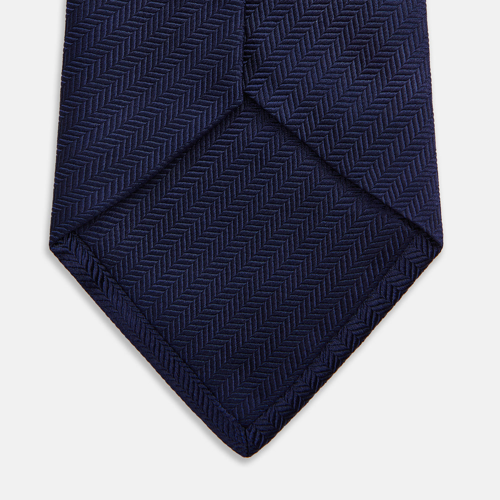 The Long Navy Herringbone Silk Tie by Turnbull & Asser is shown rolled up on a plain white background, highlighting its subtle herringbone weave and luxurious silk fabric.