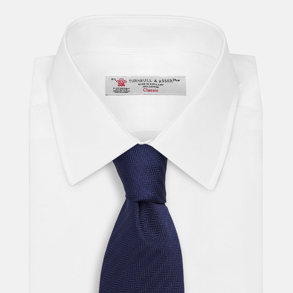 The Long Navy Herringbone Silk Tie by Turnbull & Asser is shown rolled up on a plain white background, highlighting its subtle herringbone weave and luxurious silk fabric.