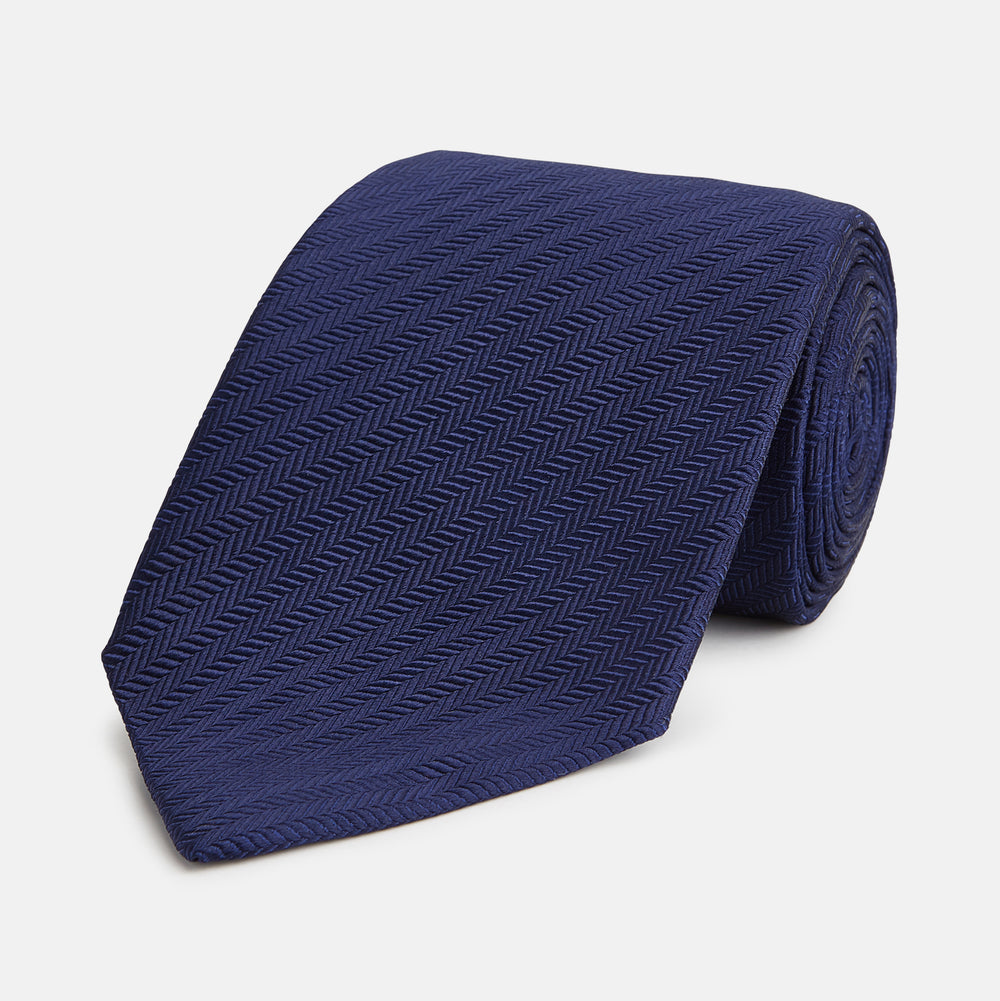 The Long Navy Herringbone Silk Tie by Turnbull & Asser is shown rolled up on a plain white background, highlighting its subtle herringbone weave and luxurious silk fabric.