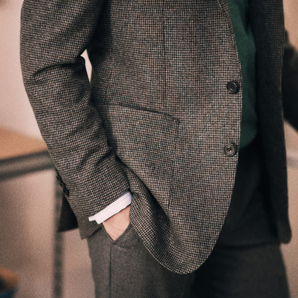 The Turnbull & Asser Charcoal Houndstooth Flannel Jacket features pure wool, a notched lapel, two front buttons, flap pockets, and buttoned cuffs, shown on a white background.