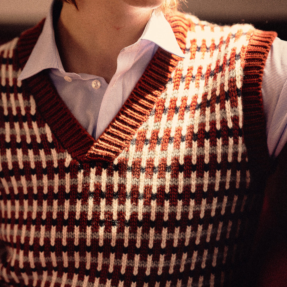 The Rust Knitted Cashmere Slipover by Turnbull & Asser is a sleeveless V-neck vest in red, white, and dark brown houndstooth, inspired by Welsh knitwear with solid red ribbed trim at the collar, armholes, and hem.