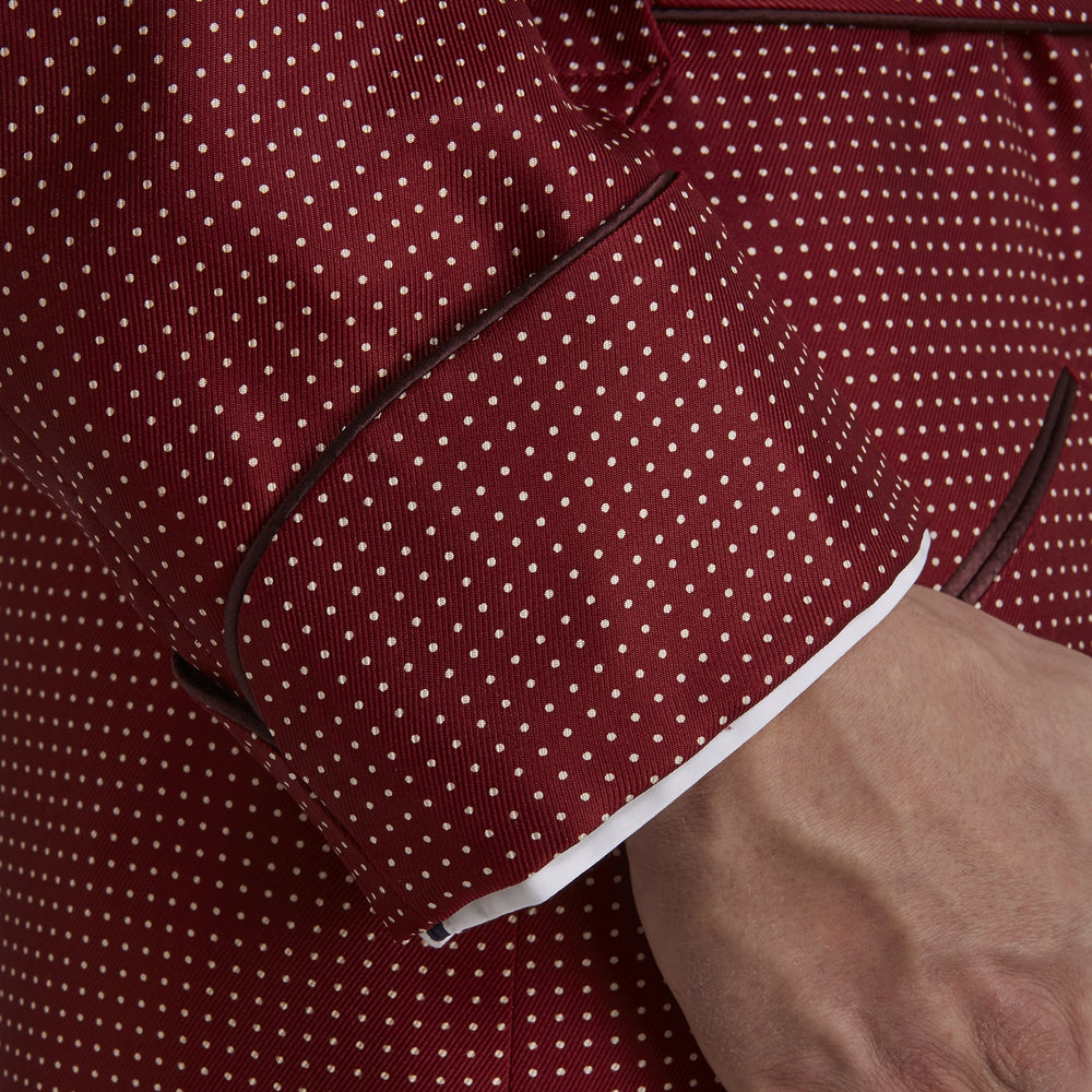 The Turnbull & Asser Burgundy Piped Silk Spot Gown features small white polka dots, a shawl collar, fabric belt, and chest pocket—luxurious nightwear and a stylish men’s statement piece—shown on a white background.