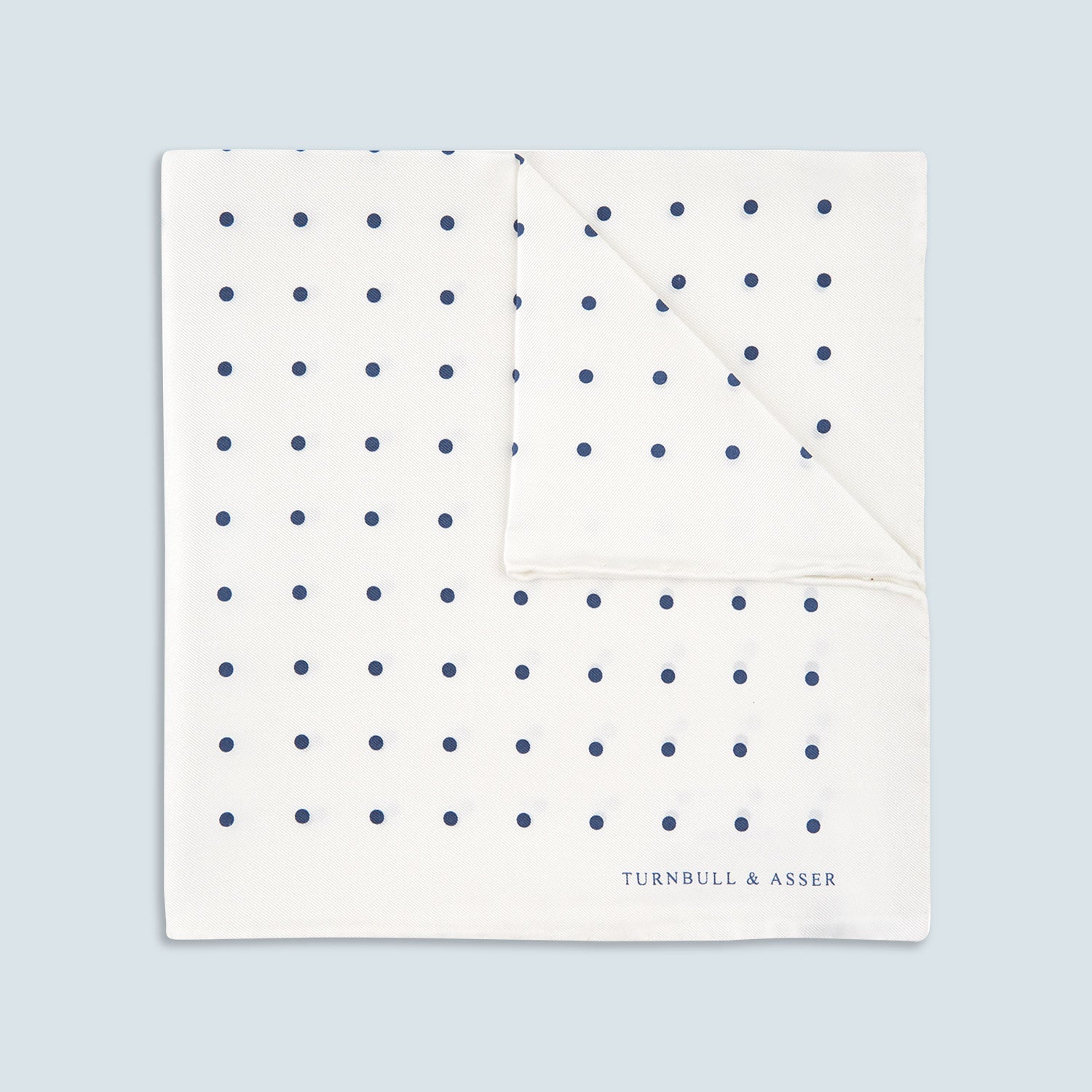 View 1 of product The White and Navy Spot Silk Pocket Square by Turnbull & Asser features evenly spaced blue dots and the brand's logo in blue on the bottom right, with a folded corner highlighting this classic men's accessory.