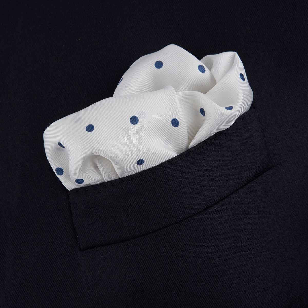 View 4 of product The White and Navy Spot Silk Pocket Square by Turnbull & Asser features evenly spaced blue dots and the brand's logo in blue on the bottom right, with a folded corner highlighting this classic men's accessory.