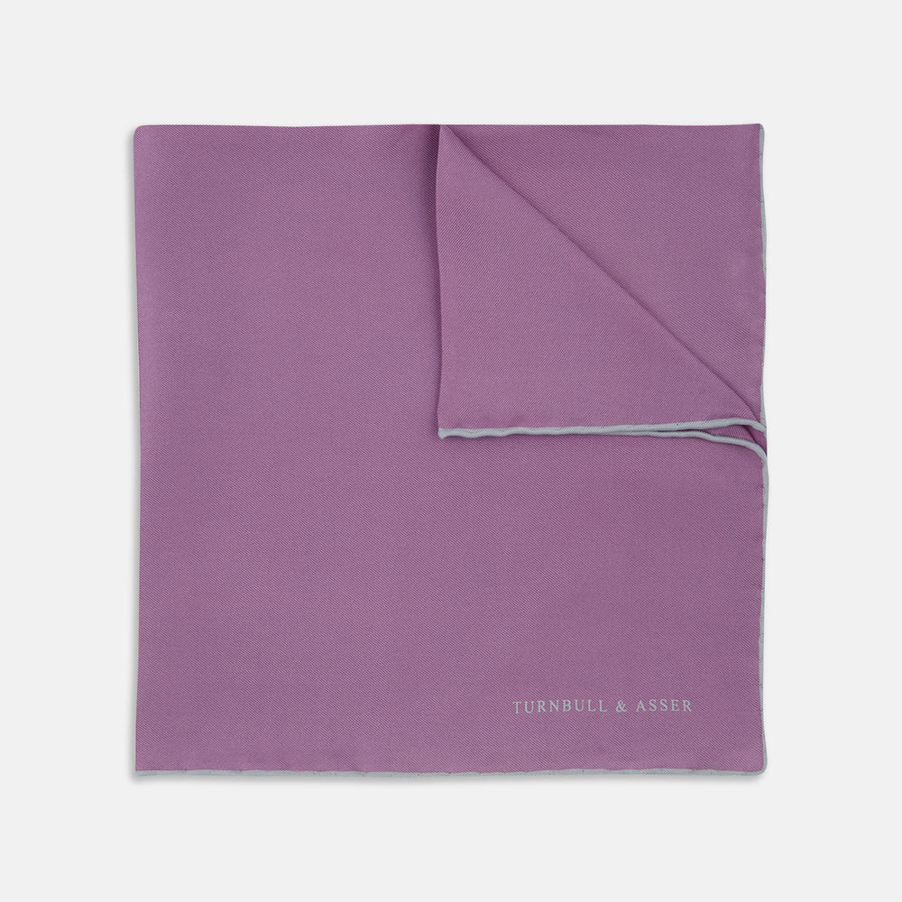The Lilac and White Piped Silk Pocket Square by Turnbull & Asser features a smooth lilac silk fabric with a refined white piped edge and the brand's signature printed in white on the lower right corner.