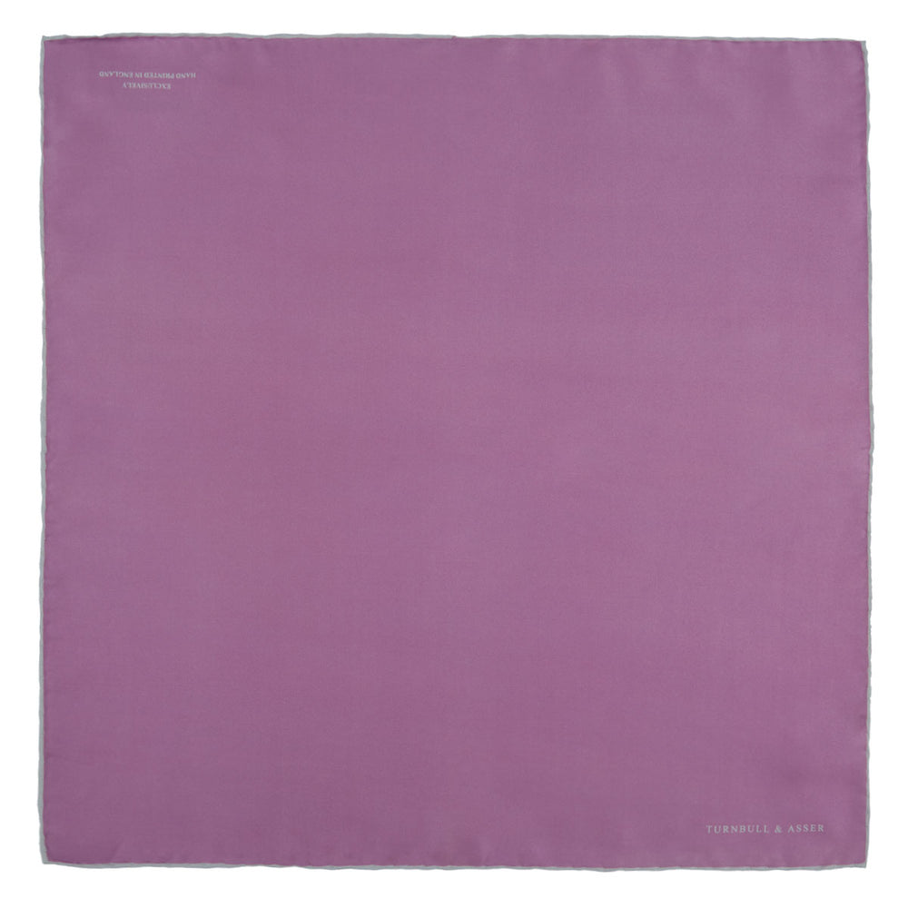 The Lilac and White Piped Silk Pocket Square by Turnbull & Asser features a smooth lilac silk fabric with a refined white piped edge and the brand's signature printed in white on the lower right corner.