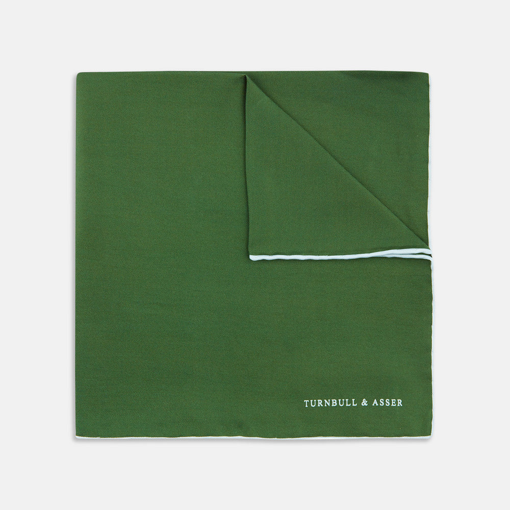 The Olive and White Piped Silk Pocket Square by Turnbull & Asser features a subtle white border and the brand’s embroidered logo—a timeless accessory for any occasion.