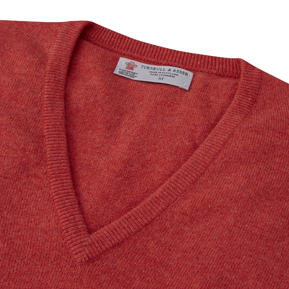 A folded Crimson Fine Cashmere Slipover by Turnbull & Asser in coral-red is shown against a white background, with the visible neckline label highlighting its premium Scottish cashmere quality.