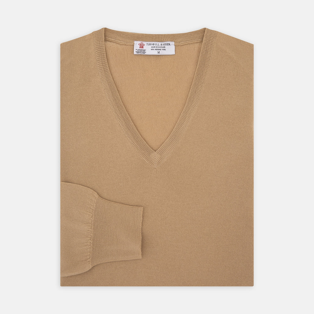 The Turnbull & Asser Camel V-Neck Merino Wool Jumper, with ribbed cuffs and a visible white neckline label, is neatly folded with one sleeve showing—an essential, versatile staple for any gentleman's wardrobe.