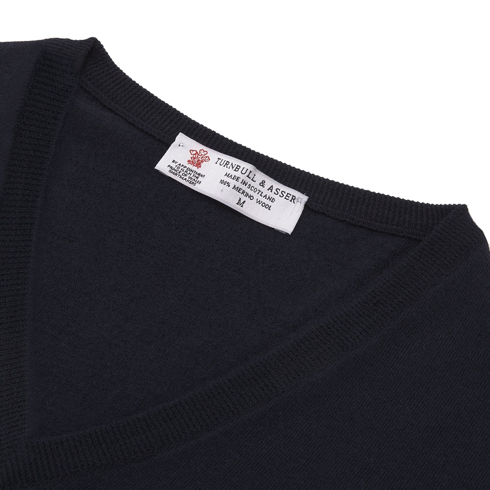 The Turnbull & Asser Indigo V-Neck Merino Wool Jumper, shown neatly folded with ribbed cuffs and a visible inner label, is a timeless essential in versatile navy, perfect for any gentleman’s wardrobe.