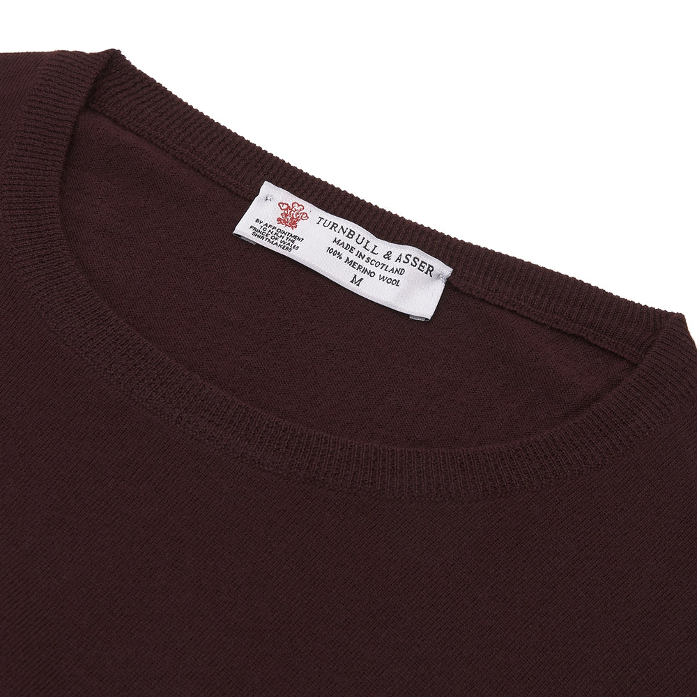 The Turnbull & Asser Wine Crew Neck Merino Wool Jumper, featuring ribbed cuffs and a classic design, is neatly folded on a white background.