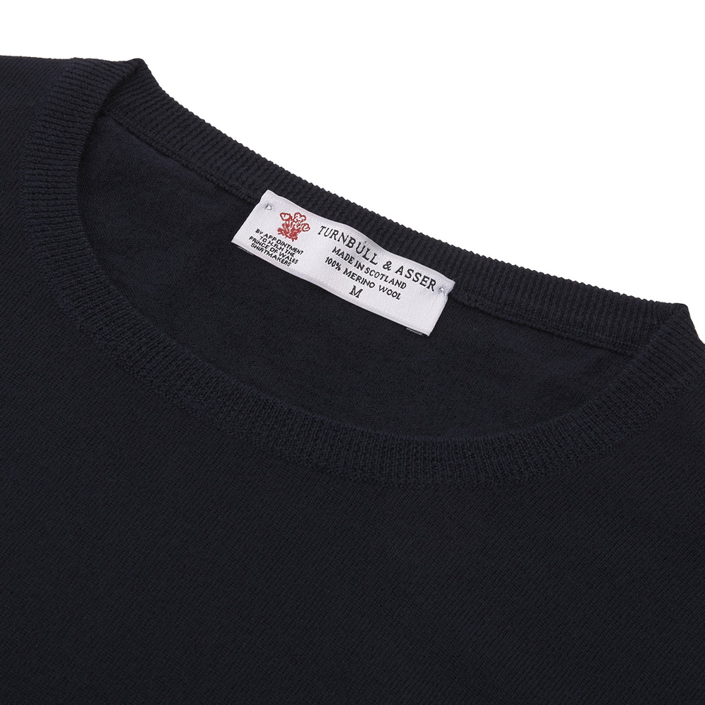 A neatly folded Indigo Crew Neck Merino Wool Jumper by Turnbull & Asser, featuring a visible brand label at the collar and one sleeve tucked across the front, displayed on a white background—a versatile staple for any gentleman's wardrobe.