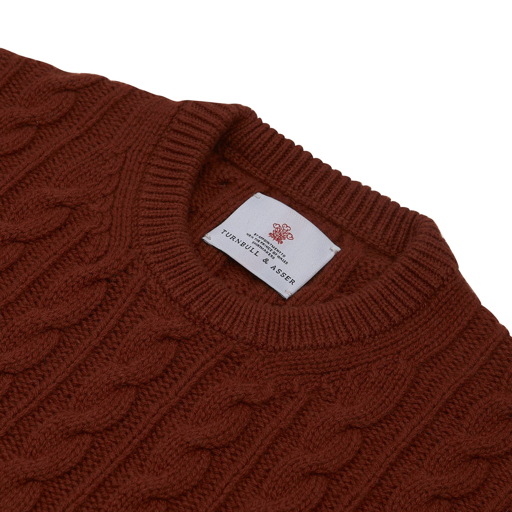 The Harissa Crew Neck Cable Knit Cashmere Jumper by Turnbull & Asser features a chunky, folded cable knit in deep harissa, with ribbed crew neckline and cuffs, and a signature label on the inside collar.