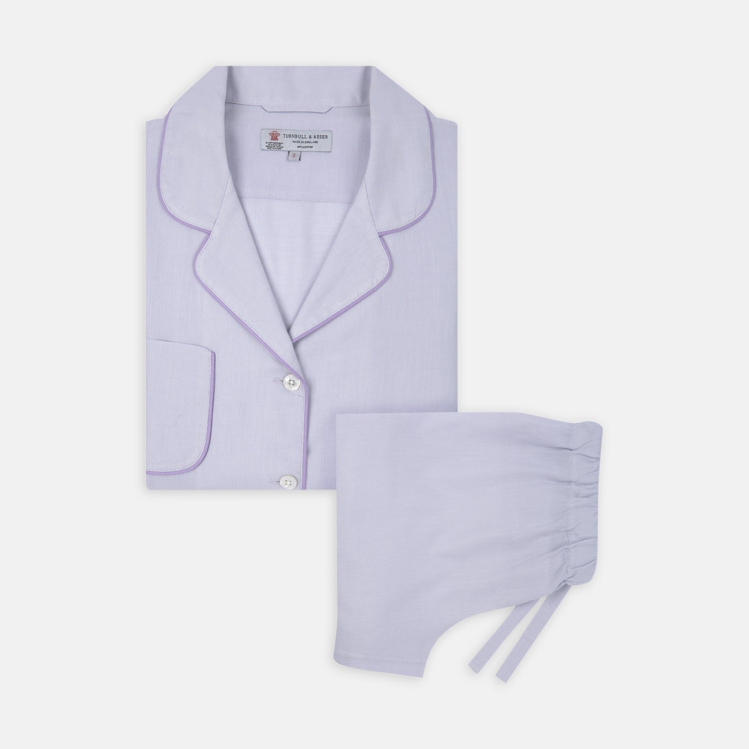 View 1 of product The Ladies' Lilac Cotton-Cashmere Pyjama Set by Turnbull & Asser, with purple piping, collared button-up top featuring a chest pocket, and matching drawstring bottoms, is folded and displayed on a white background.