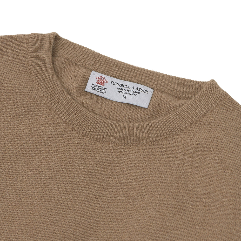 A folded Natural Crew Neck Cashmere Jumper by Turnbull & Asser, in light brown, features a visible brand label and one sleeve neatly laid across the front of this smart casual luxury Scottish sweater.