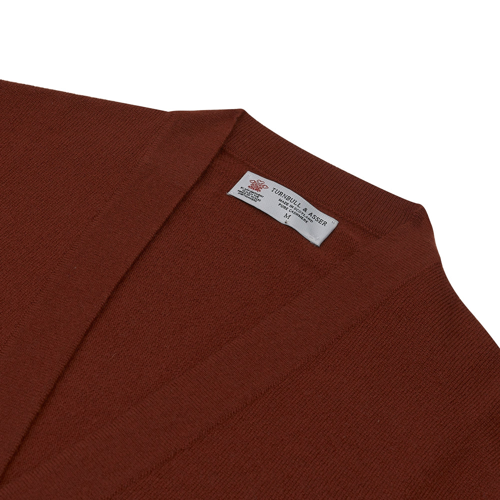 The Harissa Cashmere Cardigan by Turnbull & Asser, in deep red-brown 21GG knit, is folded to display its V-neck, ribbed cuffs, and two buttons against a white background.