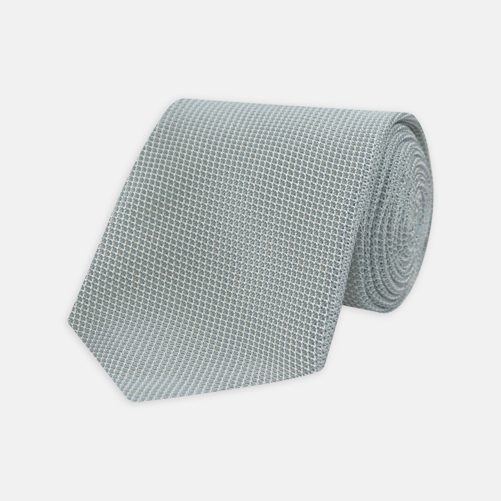 A neatly rolled Turnbull & Asser Light Blue Grenadine Silk Tie, featuring a subtle textured weave, displayed on a white background.