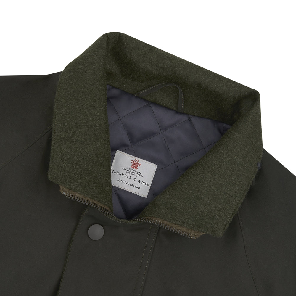 The Turnbull & Asser Dark Green Waxed Cotton Field Coat features a straight fit, button-up front, two large bottom flap pockets, slanted chest pockets, and a contrasting collar. Shown against a plain white background.