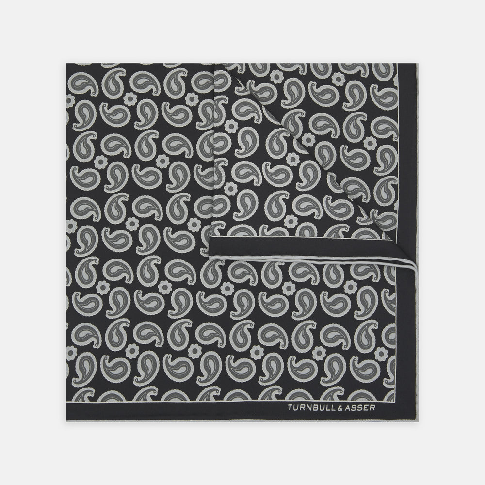 The Monochrome Paisley Mill Silk Pocket Square by Turnbull & Asser is a limited edition black silk accessory with a white paisley and floral pattern. The brand name appears in white text on the slightly folded bottom right corner.