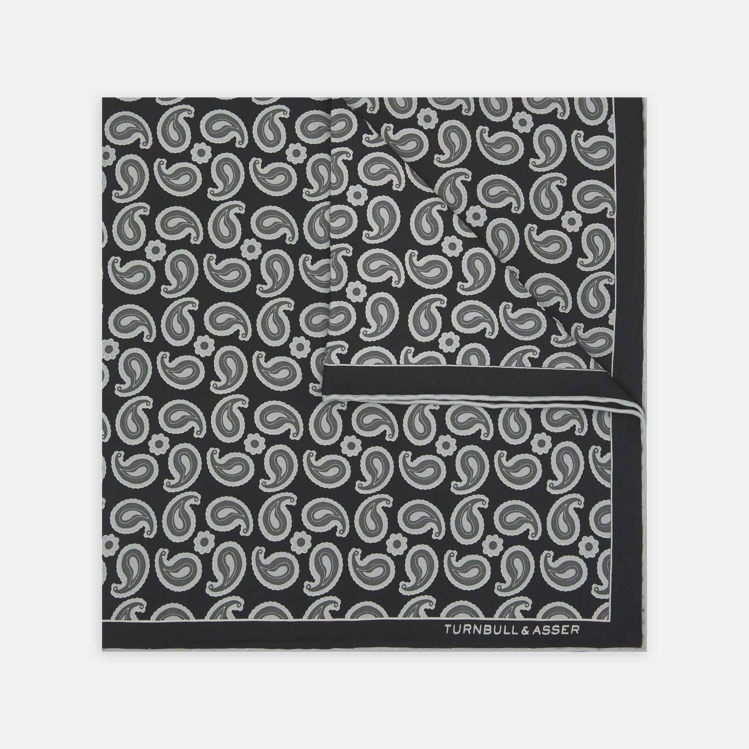 View 1 of product The Monochrome Paisley Mill Silk Pocket Square by Turnbull & Asser is a limited edition black silk accessory with a white paisley and floral pattern. The brand name appears in white text on the slightly folded bottom right corner.