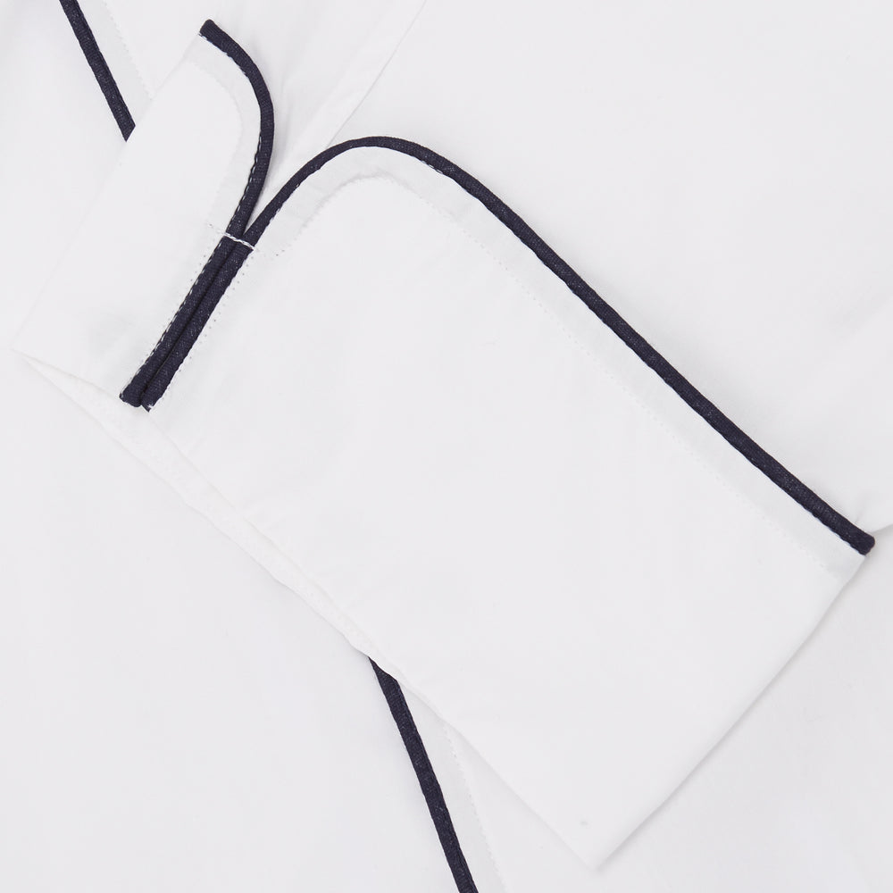 The Turnbull & Asser White Piped Cotton Pyjama Set, made from Egyptian cotton and featuring black piping and two buttons, is shown folded with a matching white tie-strap face mask on a light gray background.