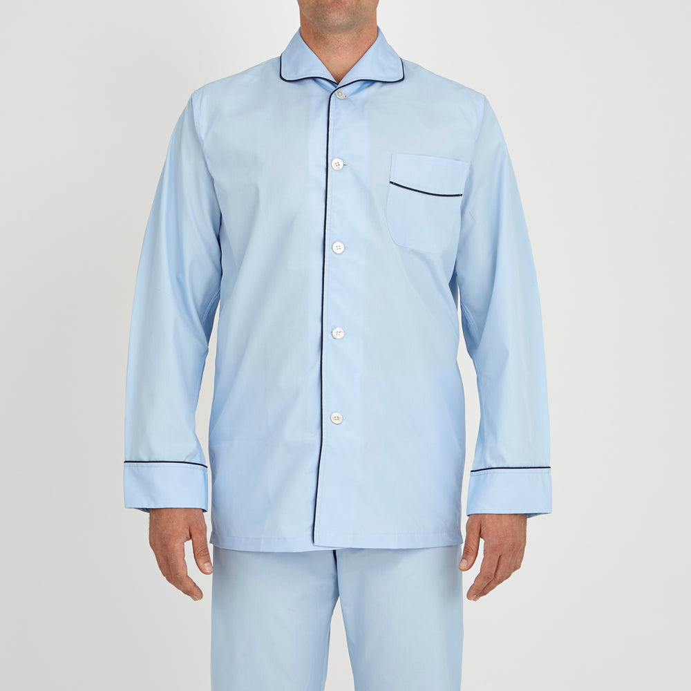 The Light Blue Piped Cotton Pyjama Set by Turnbull & Asser is neatly folded, with dark piping on the collar and buttons, displayed next to a matching light blue sleep mask with tie straps on a plain white background.