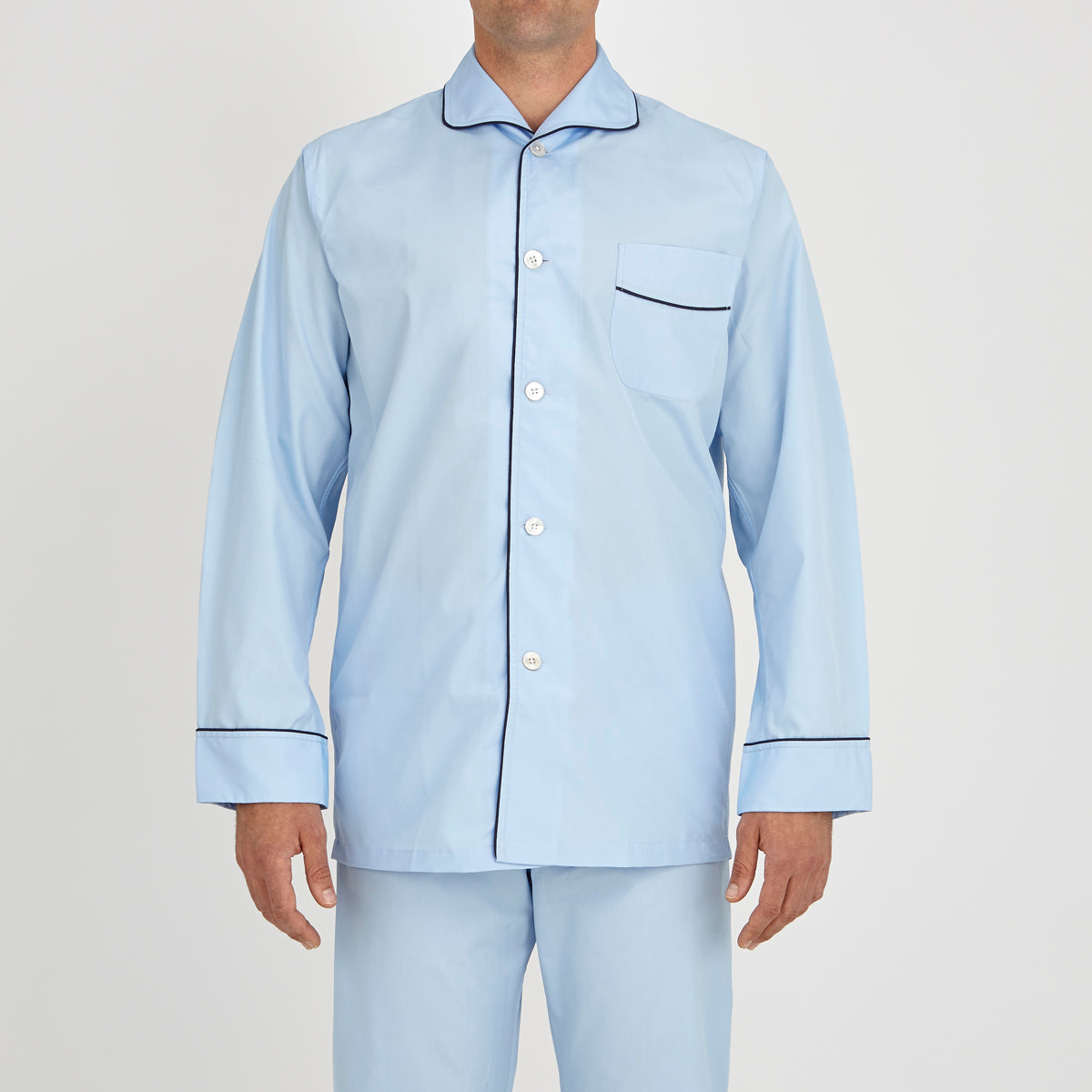 View 2 of product The Light Blue Piped Cotton Pyjama Set by Turnbull & Asser is neatly folded, with dark piping on the collar and buttons, displayed next to a matching light blue sleep mask with tie straps on a plain white background.