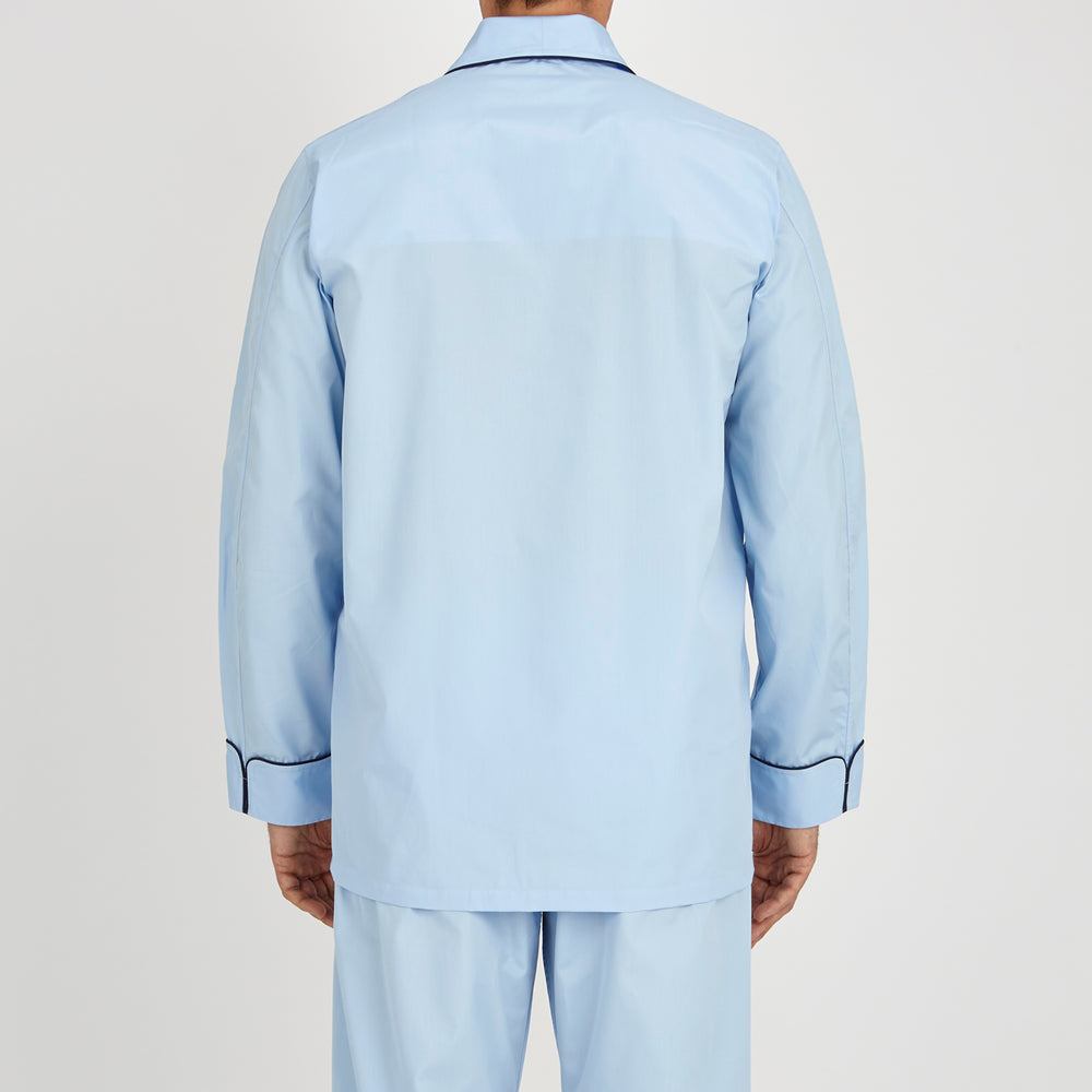 The Light Blue Piped Cotton Pyjama Set by Turnbull & Asser is neatly folded, with dark piping on the collar and buttons, displayed next to a matching light blue sleep mask with tie straps on a plain white background.