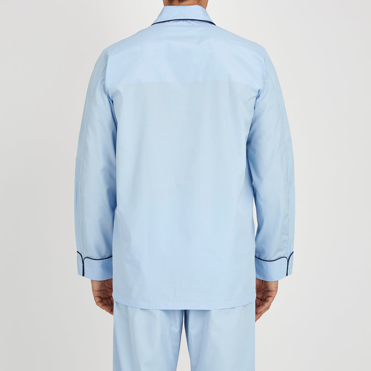 View 3 of product The Light Blue Piped Cotton Pyjama Set by Turnbull & Asser is neatly folded, with dark piping on the collar and buttons, displayed next to a matching light blue sleep mask with tie straps on a plain white background.