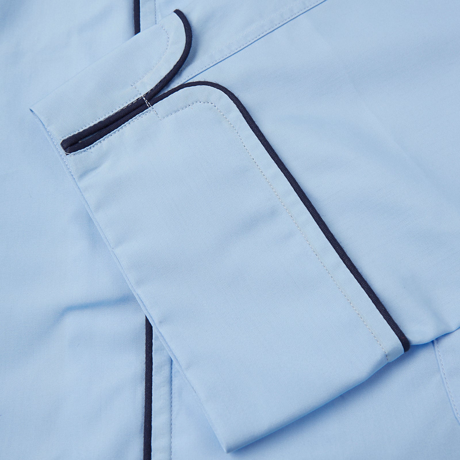 View 4 of product The Light Blue Piped Cotton Pyjama Set by Turnbull & Asser is neatly folded, with dark piping on the collar and buttons, displayed next to a matching light blue sleep mask with tie straps on a plain white background.