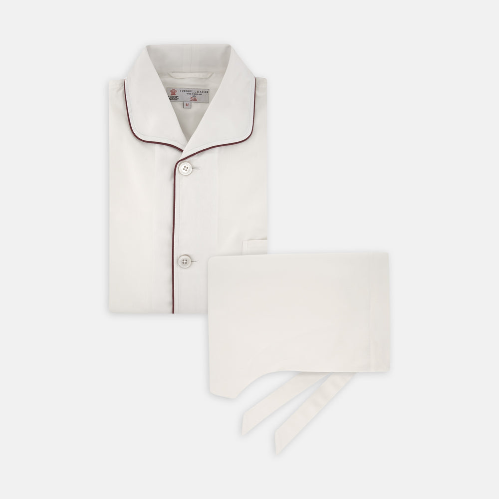The Turnbull & Asser Cream Piped Silk Pyjama Set is neatly folded with dark piping and buttons, placed next to a matching white sleep mask with tie straps, all set against a light neutral background.