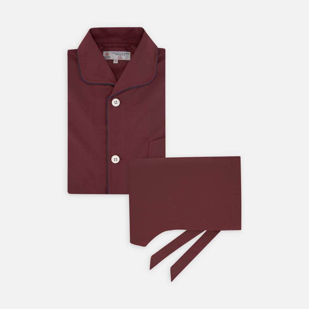 The Wine Piped Silk Pyjama Set by Turnbull & Asser features a folded maroon top with white buttons and dark trim, displayed beside a matching sleep mask with tie straps, set against a plain white background.