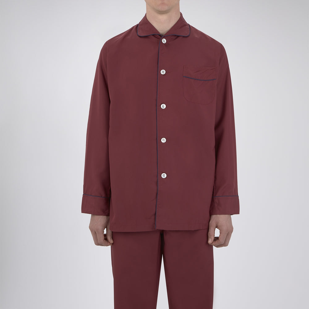 The Wine Piped Silk Pyjama Set by Turnbull & Asser features a folded maroon top with white buttons and dark trim, displayed beside a matching sleep mask with tie straps, set against a plain white background.