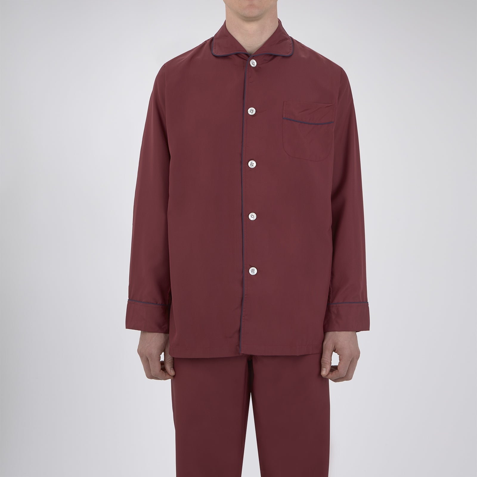 View 2 of product The Wine Piped Silk Pyjama Set by Turnbull & Asser features a folded maroon top with white buttons and dark trim, displayed beside a matching sleep mask with tie straps, set against a plain white background.