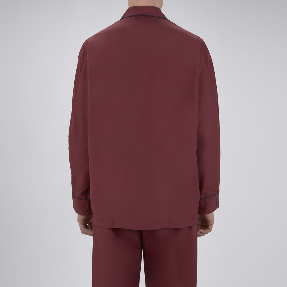 The Wine Piped Silk Pyjama Set by Turnbull & Asser features a folded maroon top with white buttons and dark trim, displayed beside a matching sleep mask with tie straps, set against a plain white background.