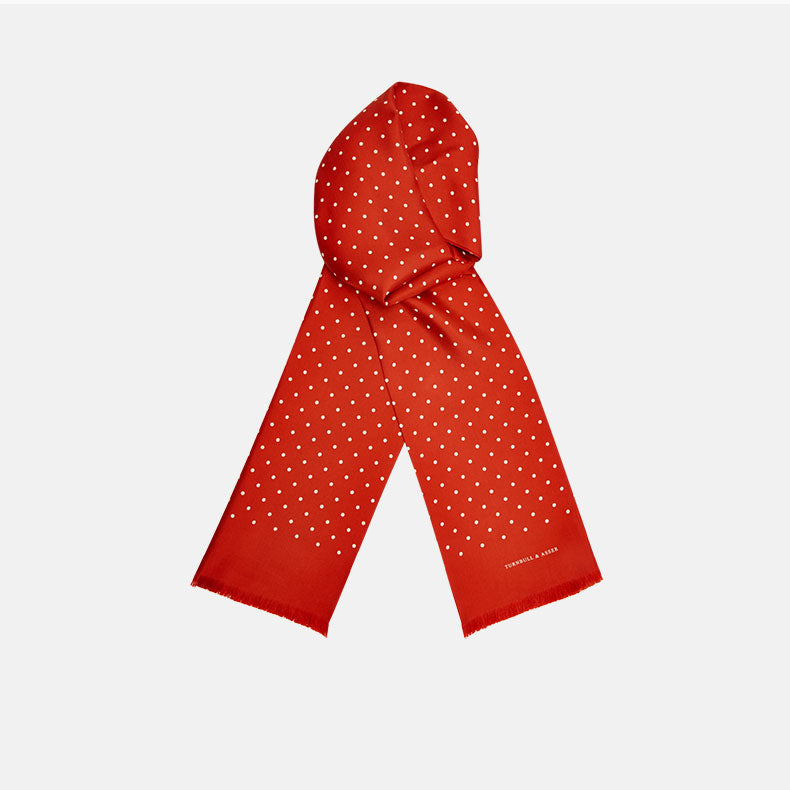 The Red and White Spotted Silk Scarf by Turnbull & Asser features white polka dots and fringed edges on silk. The brand name appears subtly on one end, with the scarf displayed against a plain light background.