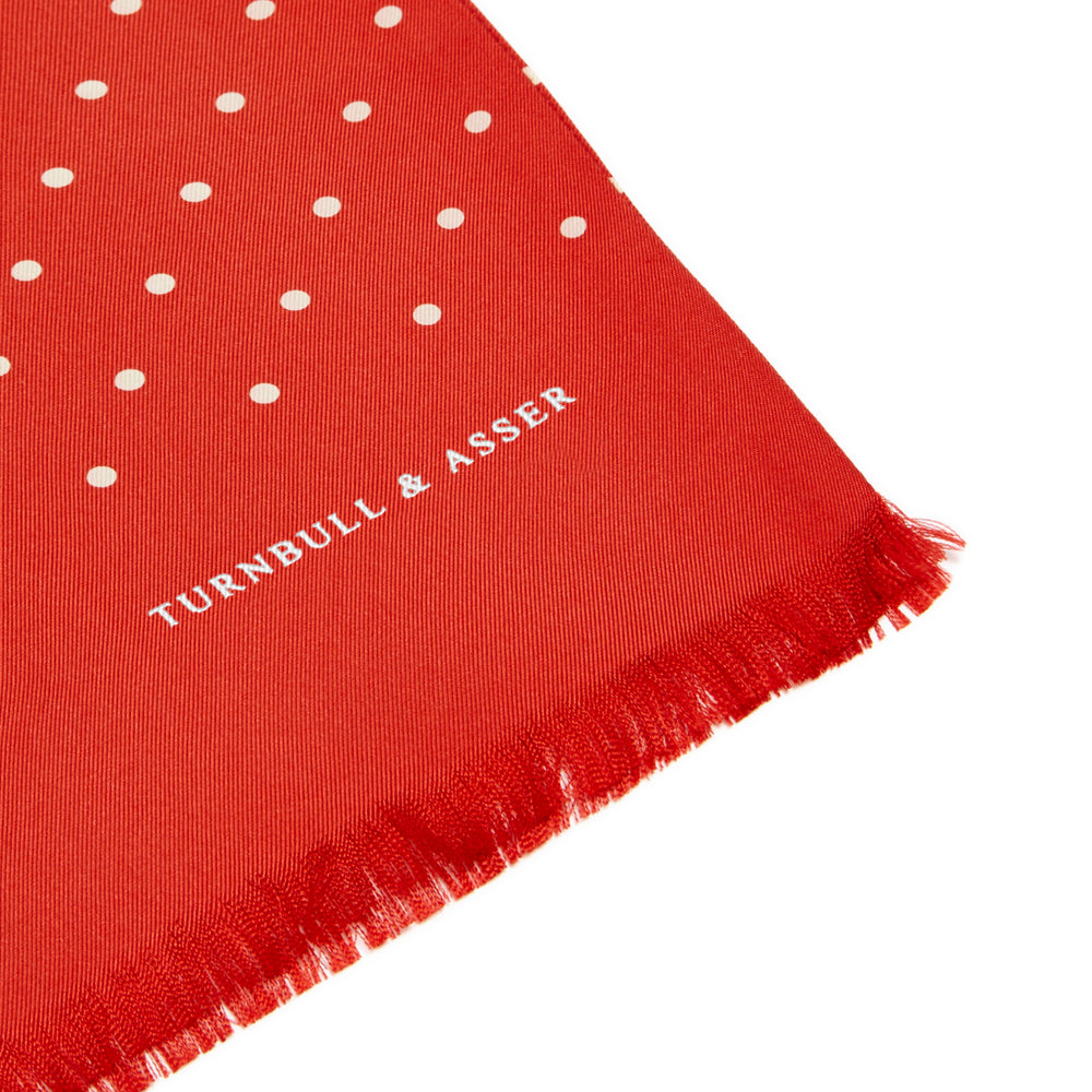 The Red and White Spotted Silk Scarf by Turnbull & Asser features white polka dots and fringed edges on silk. The brand name appears subtly on one end, with the scarf displayed against a plain light background.
