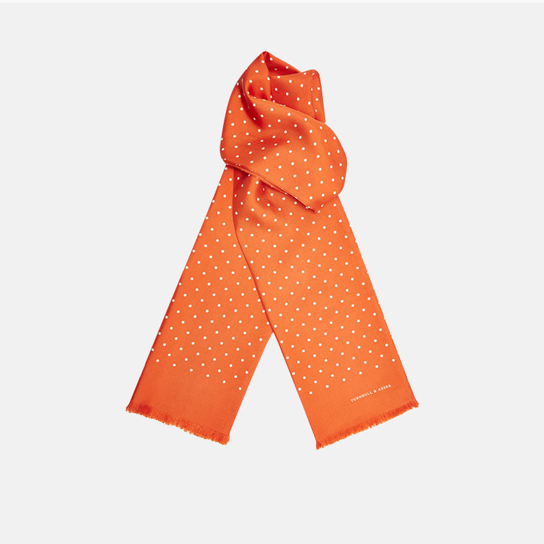 The Turnbull & Asser Orange and White Spotted Silk Scarf, featuring white polka dots and fringed edges, is neatly folded and displayed on a plain light gray background.