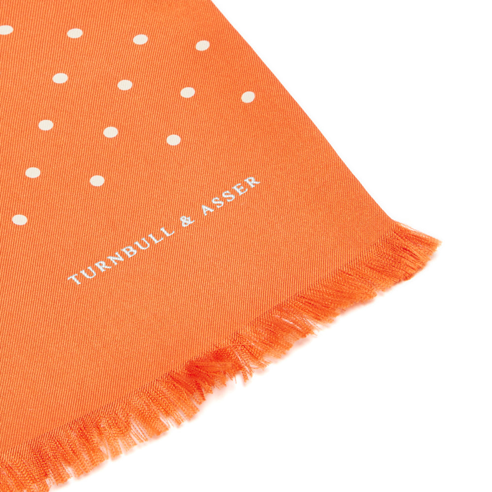 The Turnbull & Asser Orange and White Spotted Silk Scarf, featuring white polka dots and fringed edges, is neatly folded and displayed on a plain light gray background.